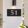 Brushed silver house number sign with a brushed metal look, The Kawana, black face with the number 51 in silver lettering, on white weatherboards under a black wall light, beside a timber front door.