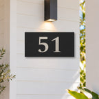 Brushed silver house number sign with a brushed metal look, The Kawana, black face with the number 51 in silver lettering, on white weatherboards under a black wall light, beside a timber front door.