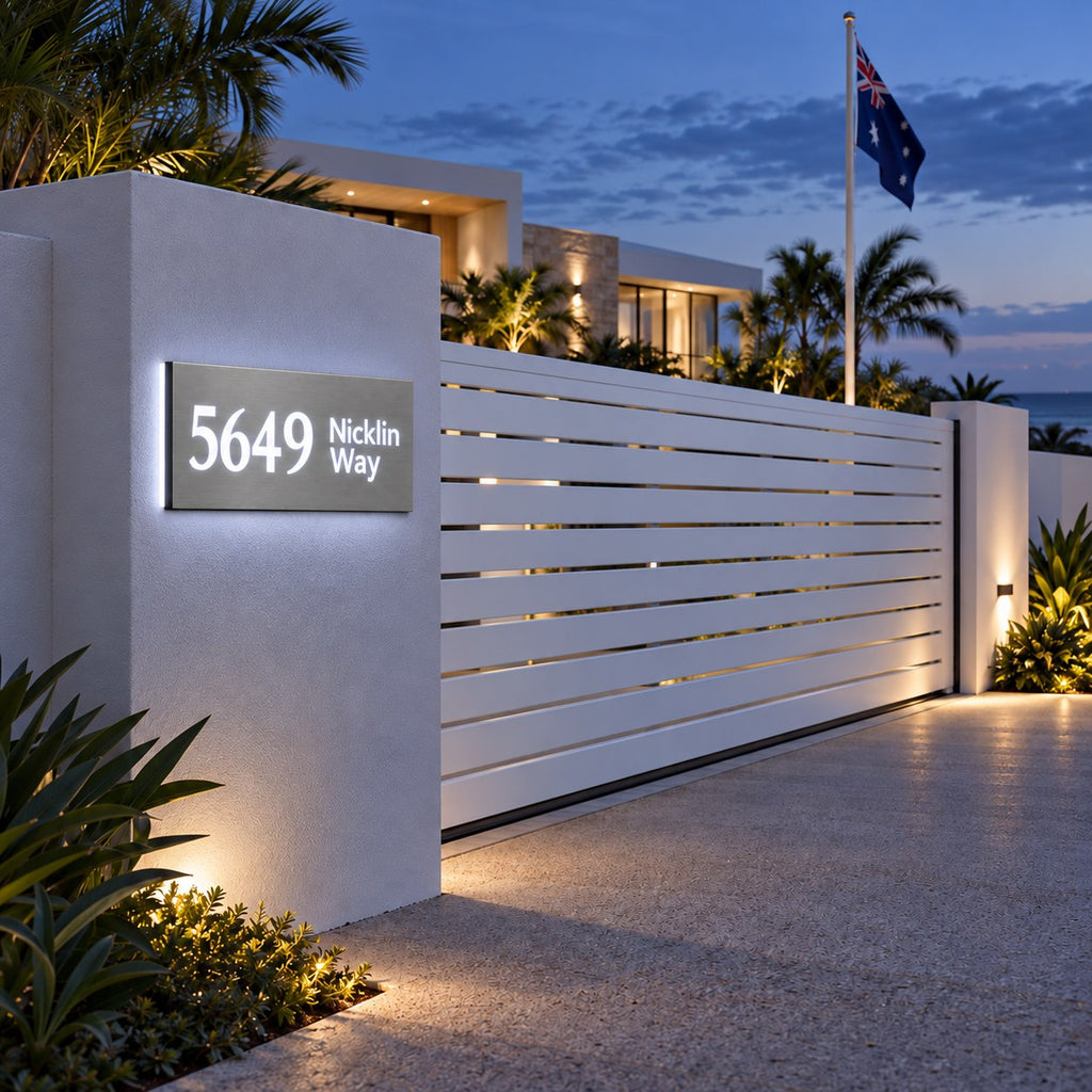 Brushed silver illuminated house number and street name sign with a brushed metal look, The Marcoola, mounted on a white rendered entry pillar beside a slat gate at dusk, 5649 Nicklin Way lit cool white.