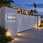 Brushed silver illuminated house number and street name sign with a brushed metal look, The Marcoola, mounted on a white rendered entry pillar beside a slat gate at dusk, 5649 Nicklin Way lit cool white.