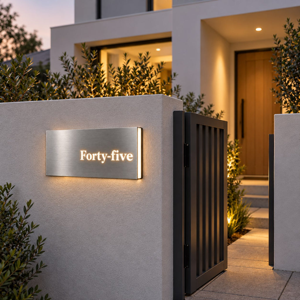 Brushed silver illuminated house number sign with a brushed metal look, The Montville, mounted on a rendered entry wall beside the gate of a white home at dusk, the number spelled out as Forty-five with the light spilling around it.