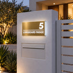 Brushed gold illuminated house number and street name sign with a brushed metal look, The Noosa, mounted on a white rendered entry pillar above a letterbox beside a slat gate at dusk, 5 Longboard Parade lit cool white.