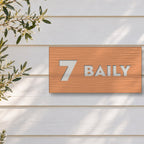 Brushed copper house number and street name sign with a brushed metal look, The Maleny, reading 7 Baily in white lettering, on white weatherboards beside an olive tree.