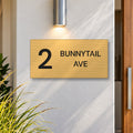 Brushed gold house number and street name sign with a brushed metal look, The Maleny, reading 2 Bunnytail Ave in black lettering, on a white rendered wall under a wall light, beside a timber front door.