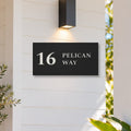 Matte black house number and street name sign, The Maleny, reading 16 Pelican Way in white lettering, on white weatherboards under a black wall light, beside a timber front door.