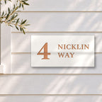 Brushed copper house number and street name sign with a brushed metal look, The Marcoola, white face reading 4 Nicklin Way in copper lettering, on white weatherboards beside a black-framed window and an olive tree.