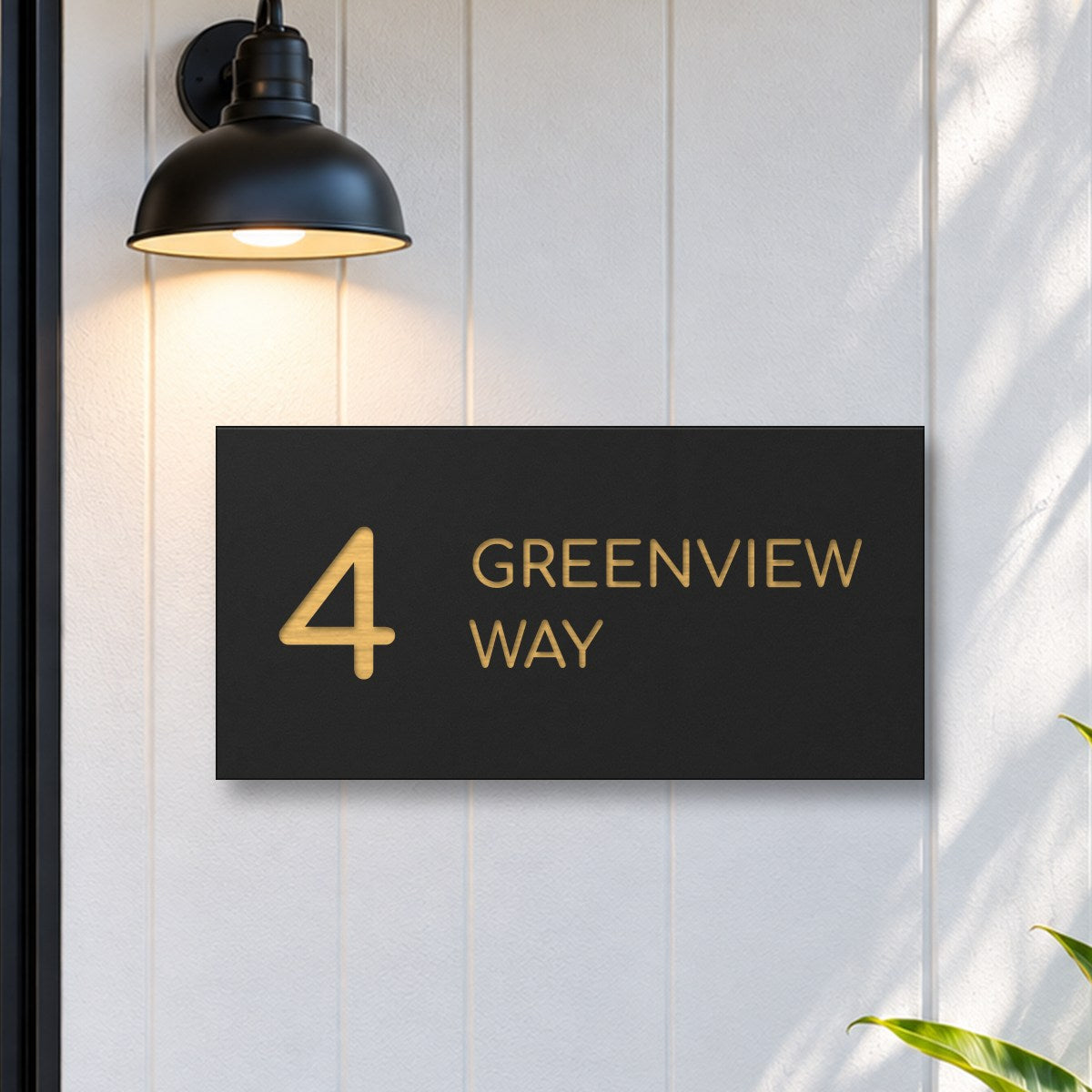 Brushed gold house number and street name sign with a brushed metal look, The Marcoola, black face reading 4 Greenview Way in gold lettering, on white cladding under a black wall light, beside a black front door.