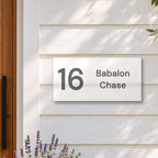 Matte white house number and street name sign, The Marcoola, reading 16 Babalon Chase in charcoal lettering, on white weatherboards beside a timber front door, with lavender and an olive tree.