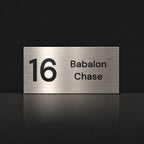 Brushed silver house number and street name sign with a brushed metal look, The Marcoola, reading 16 Babalon Chase in black lettering.