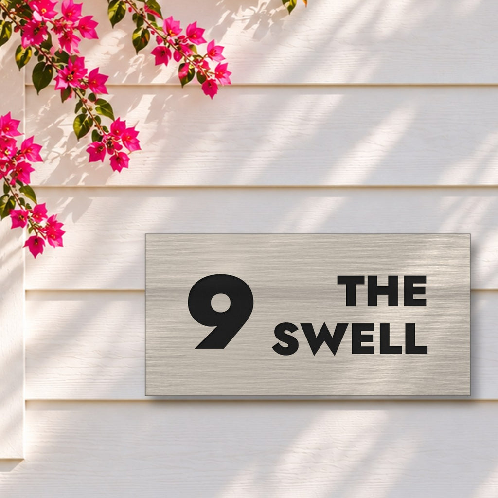 Brushed silver house number and street name sign with a brushed metal look, The Marcoola, reading 9 The Swell in black lettering, on white weatherboards under a pink bougainvillea.