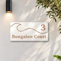 Brushed copper house number and street name sign with a brushed metal look, The Moffat, white face reading 3 Bungalow Court in copper lettering, with a wave, on a white rendered wall under a black wall light, beside an olive tree.