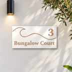 Brushed copper house number and street name sign with a brushed metal look, The Moffat, white face reading 3 Bungalow Court in copper lettering, with a wave, on a white rendered wall under a black wall light, beside an olive tree.