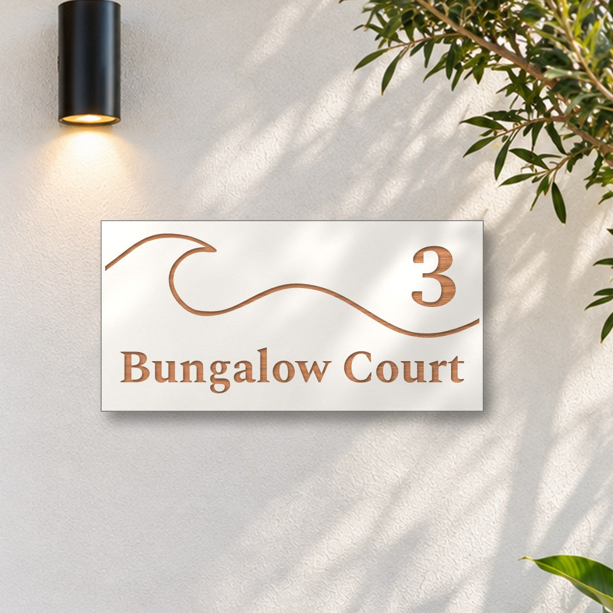 Brushed copper house number and street name sign with a brushed metal look, The Moffat, white face reading 3 Bungalow Court in copper lettering, with a wave, on a white rendered wall under a black wall light, beside an olive tree.