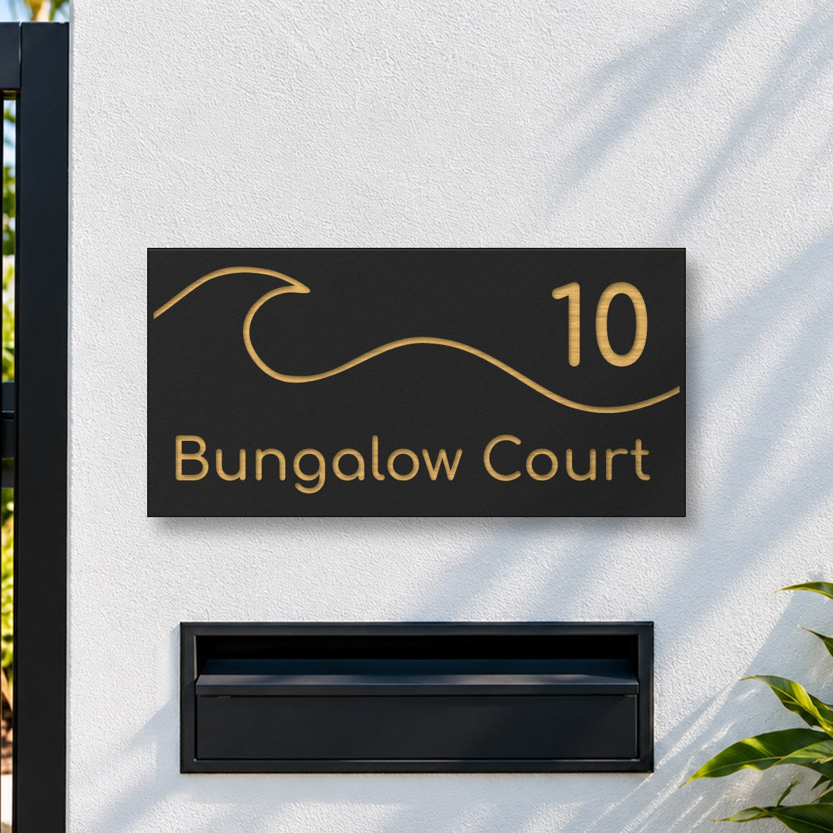 Brushed gold house number and street name sign with a brushed metal look, The Moffat, black face reading 10 Bungalow Court in gold lettering, with a wave, on a white rendered fence above a black letterbox.