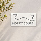 Matte white house number and street name sign, The Moffat, reading 7 Moffat Court in charcoal lettering, with a wave, on a white rendered wall with olive branches.