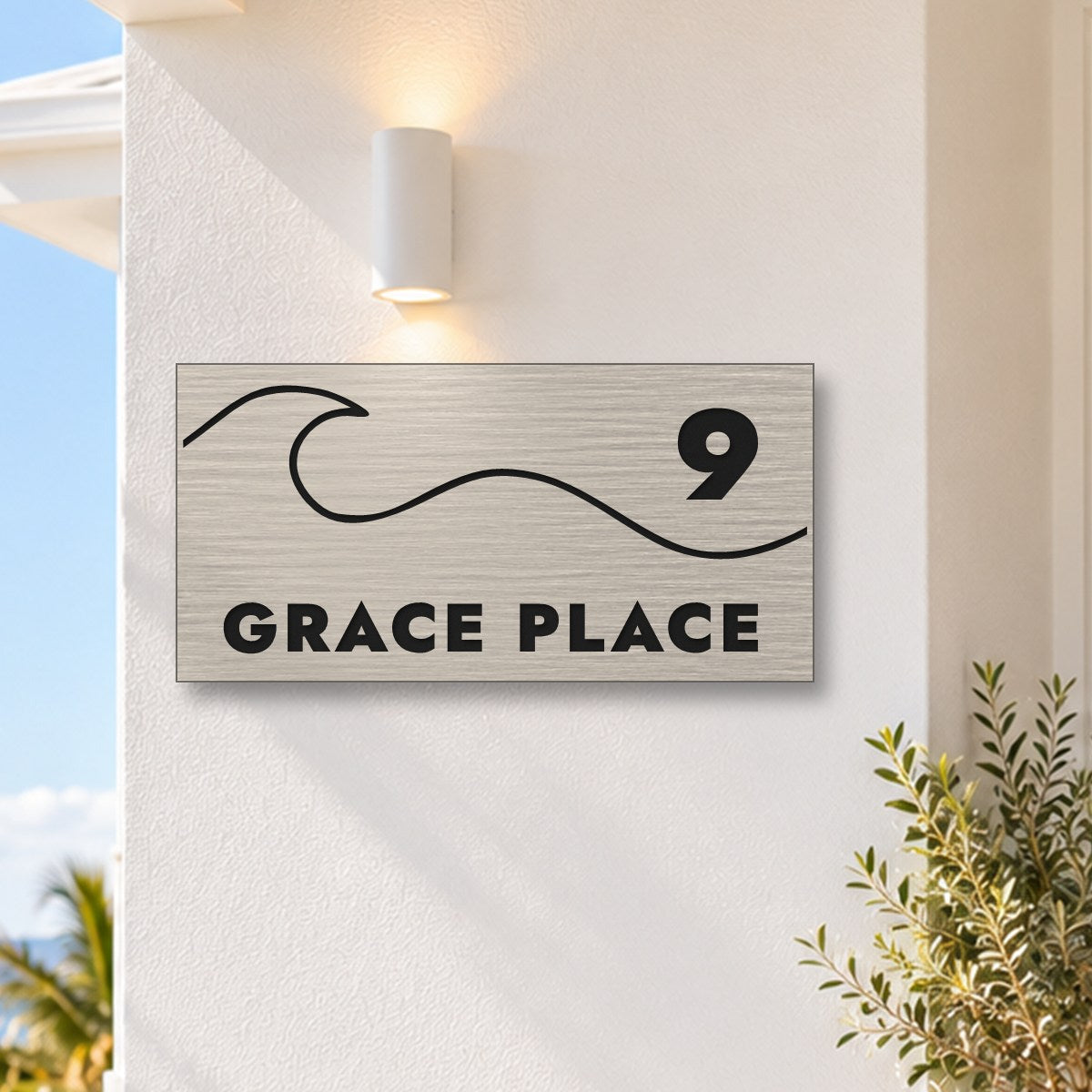Brushed silver house number and street name sign with a brushed metal look, The Moffat, reading 9 Grace Place in black lettering, with a wave, on a white rendered wall under a wall light, with the ocean behind.