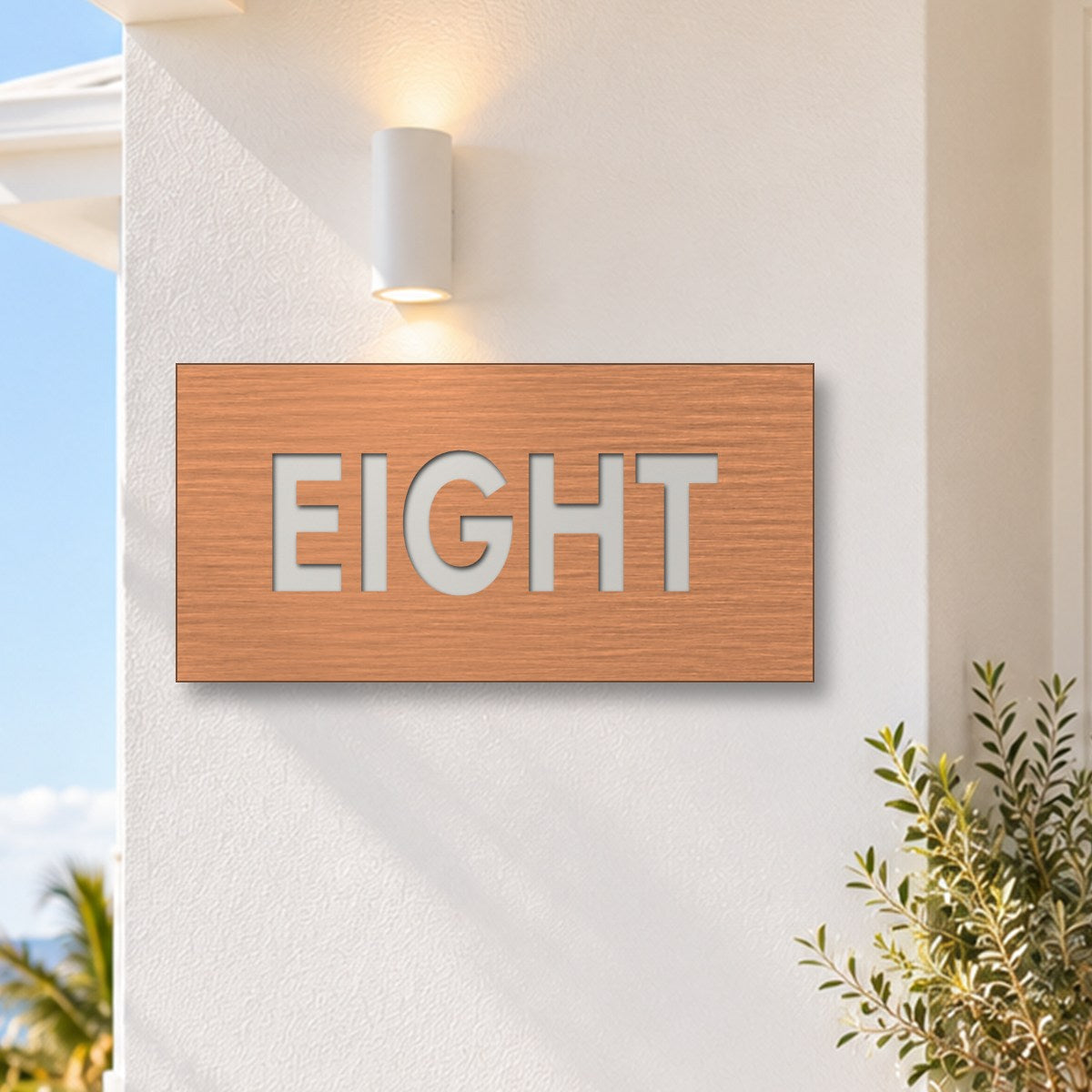 Brushed copper house number sign with a brushed metal look, The Montville, with the number spelled out as eight in white lettering, on a white rendered wall under a wall light, with the ocean behind.