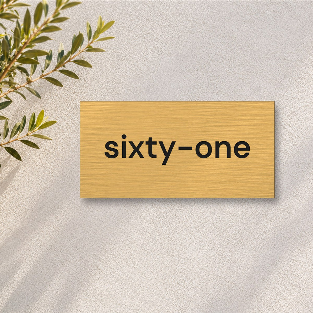 Brushed gold house number sign with a brushed metal look, The Montville, with the number spelled out as sixty-one in black lettering, on a white rendered wall with olive branches.
