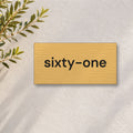 Brushed gold house number sign with a brushed metal look, The Montville, with the number spelled out as sixty-one in black lettering, on a white rendered wall with olive branches.