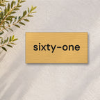 Brushed gold house number sign with a brushed metal look, The Montville, with the number spelled out as sixty-one in black lettering, on a white rendered wall with olive branches.