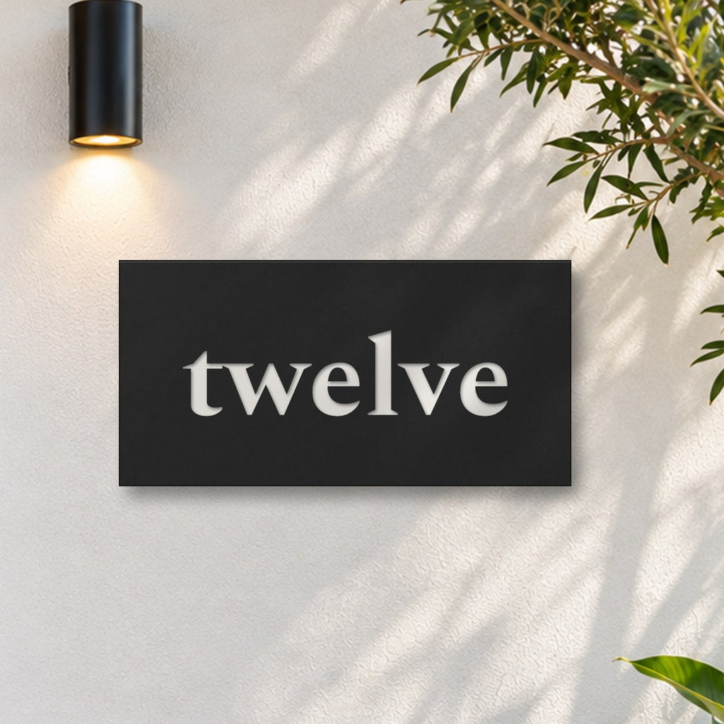 Matte black house number sign, The Montville, with the number spelled out as twelve in white lettering, on a white rendered wall under a black wall light, beside an olive tree.