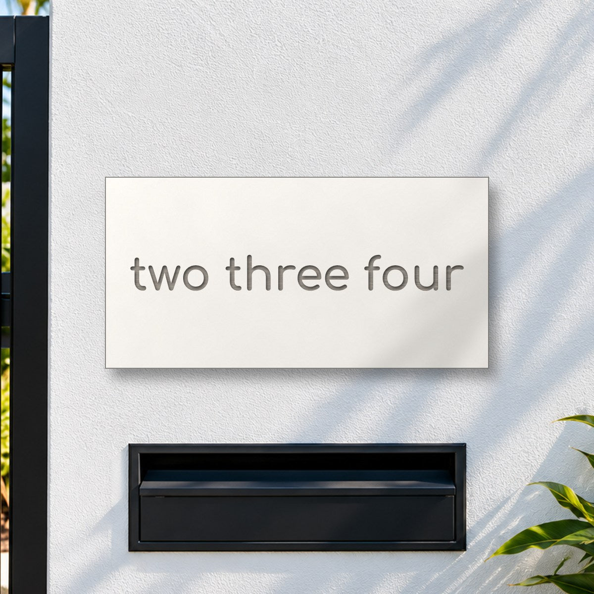Brushed silver house number sign with a brushed metal look, The Montville, white face with the number spelled out as two three four in silver lettering, on a white rendered fence above a black letterbox.