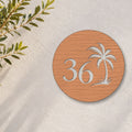 Brushed copper round house number sign with the number 36 and a palm tree cut through to white, The Mooloolaba style, on a white rendered wall with olive branches.