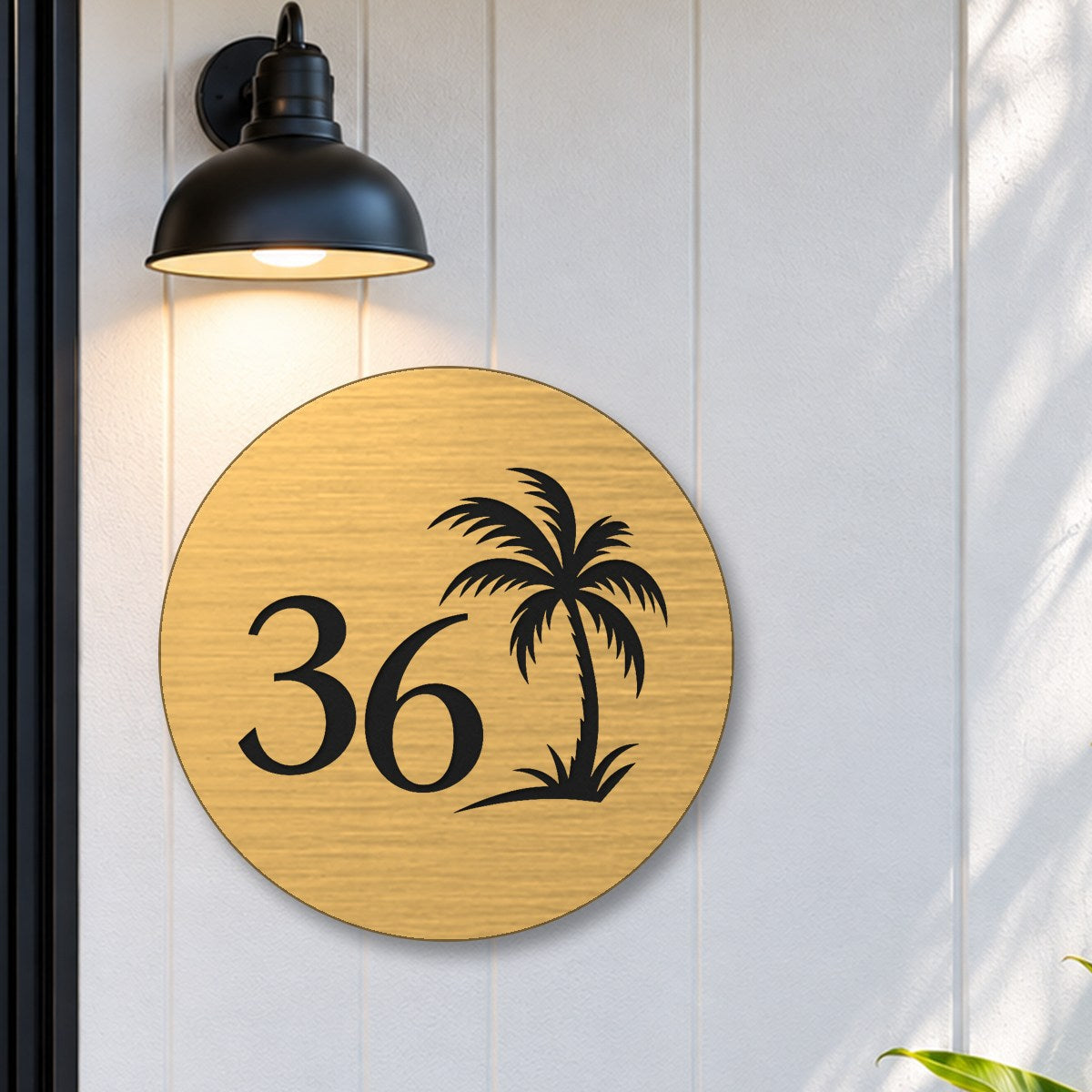 Brushed gold round house number sign with the number 36 and a palm tree cut through to black, The Mooloolaba style, on white cladding under a black wall light, beside a black front door.