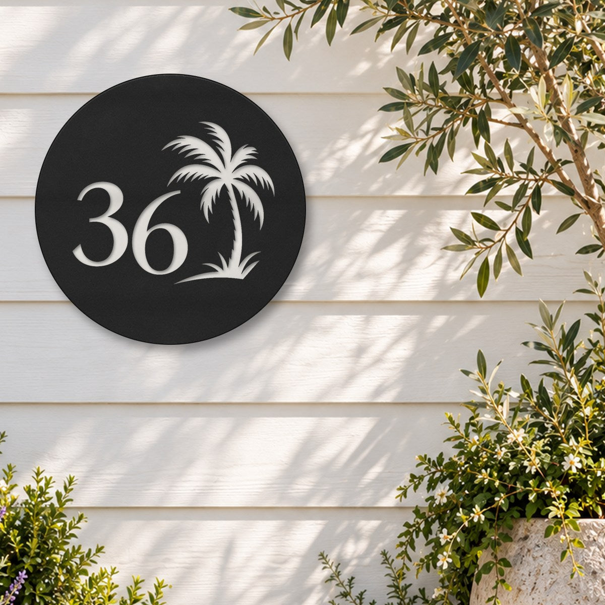Matte black round house number sign with the number 36 and a palm tree cut through to white, The Mooloolaba style, on white weatherboards beside an olive tree and flowering shrubs.