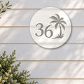 White round house number sign with the number 36 and a palm tree cut through to brushed silver, The Mooloolaba style, on white weatherboards above an olive tree in a stone pot.