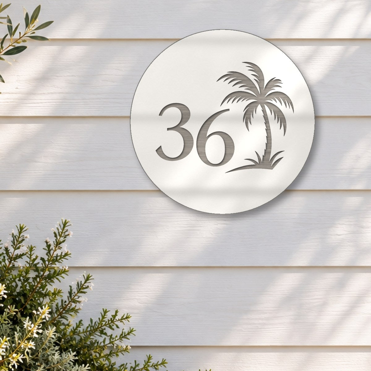 White round house number sign with the number 36 and a palm tree cut through to brushed silver, The Mooloolaba style, on white weatherboards above an olive tree in a stone pot.