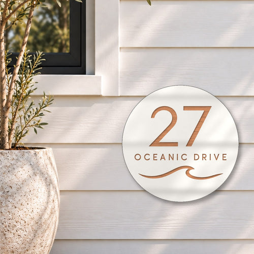 White round house number and street name sign reading 27 Oceanic Drive with a curling wave cut through to brushed copper, The Mudjimba style, on white weatherboards below a black-framed window, beside an olive tree.
