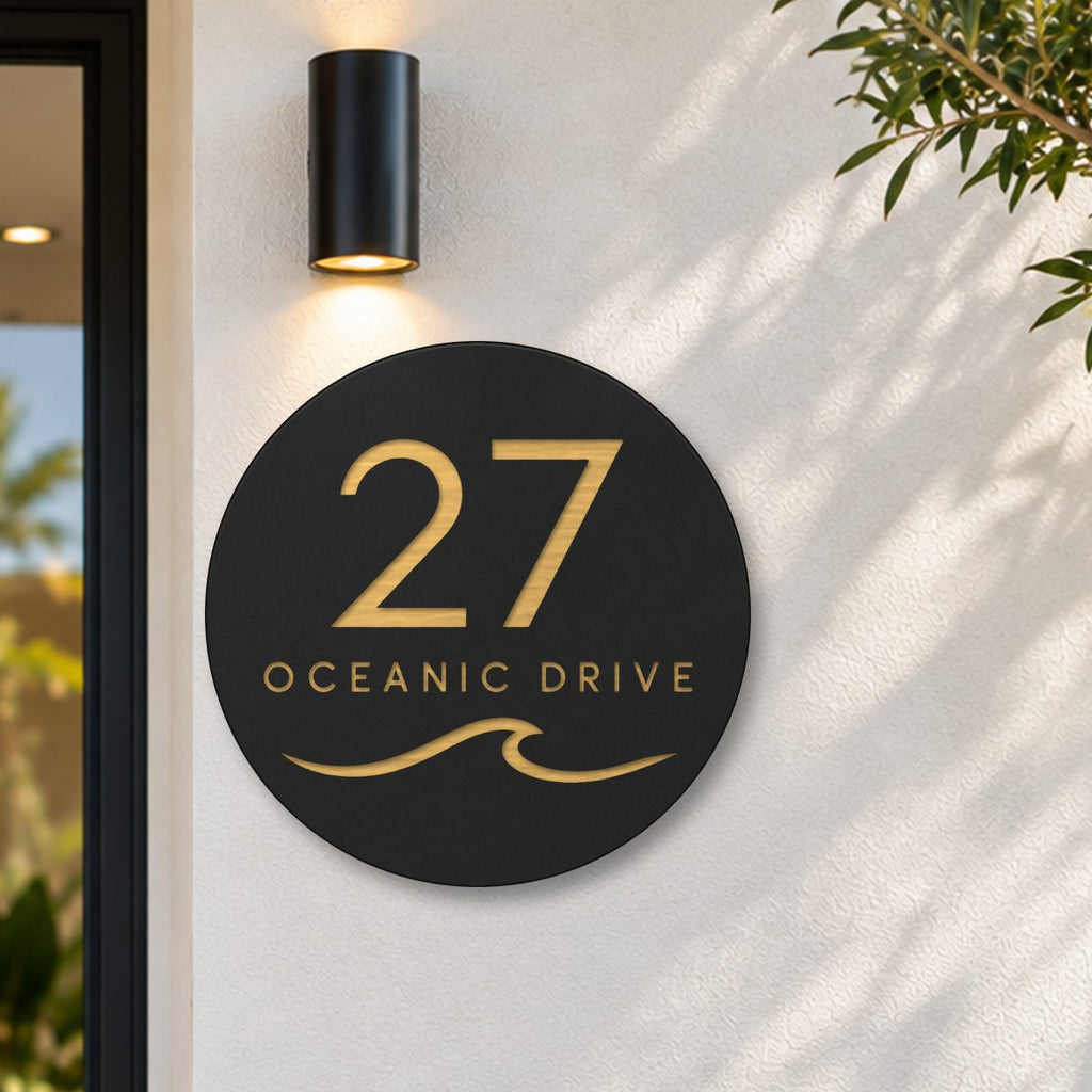Black round house number and street name sign reading 27 Oceanic Drive with a curling wave cut through to brushed gold, The Mudjimba style, on a white rendered wall under a black wall light, beside the front door.