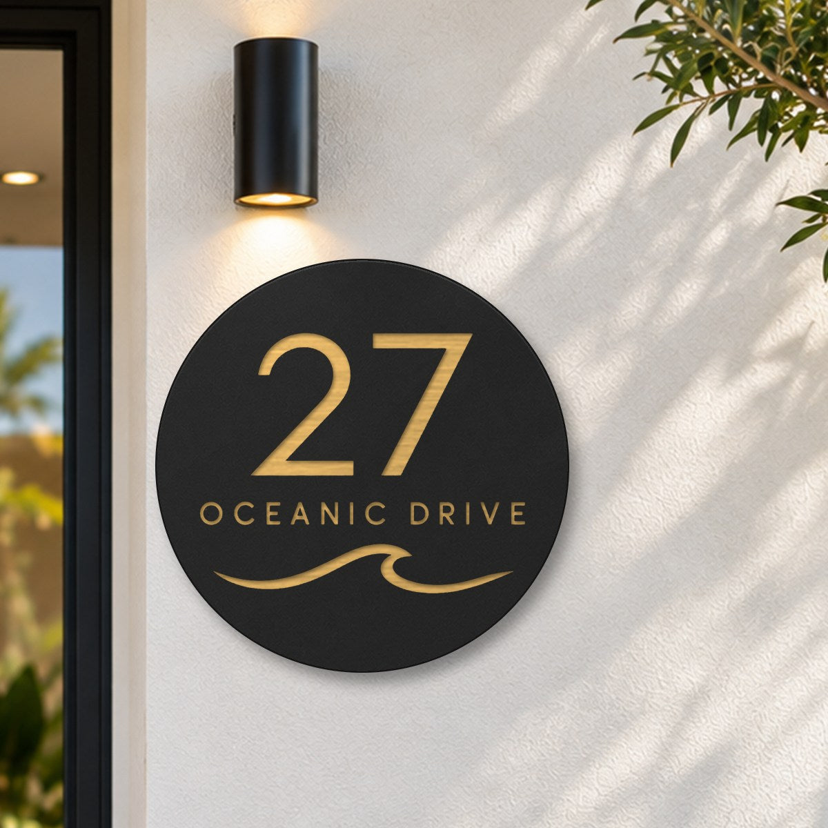 Black round house number and street name sign reading 27 Oceanic Drive with a curling wave cut through to brushed gold, The Mudjimba style, on a white rendered wall under a black wall light, beside the front door.