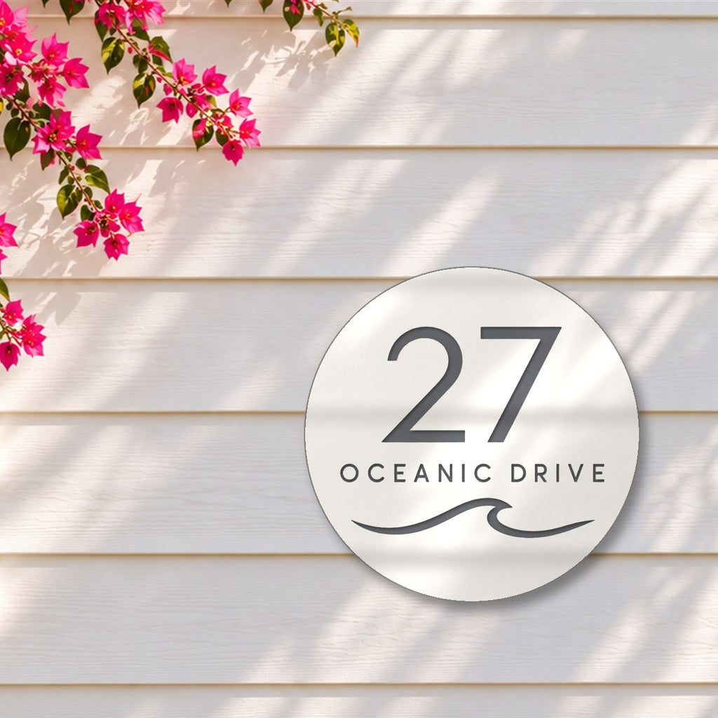 Matte white round house number and street name sign reading 27 Oceanic Drive with a curling wave cut through to charcoal, The Mudjimba style, on white weatherboards under a pink bougainvillea.