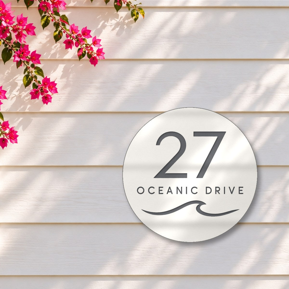 Matte white round house number and street name sign reading 27 Oceanic Drive with a curling wave cut through to charcoal, The Mudjimba style, on white weatherboards under a pink bougainvillea.