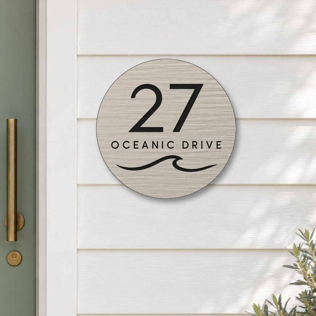 Brushed silver round house number and street name sign reading 27 Oceanic Drive with a curling wave cut through to black, The Mudjimba style, on white weatherboards beside a sage green front door.