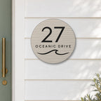 Brushed silver round house number and street name sign reading 27 Oceanic Drive with a curling wave cut through to black, The Mudjimba style, on white weatherboards beside a sage green front door.