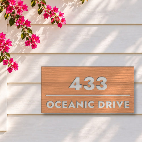 Brushed copper house number and street name sign with a brushed metal look, The Noosa, reading 433 Oceanic Drive in white lettering, on white weatherboards under a pink bougainvillea.