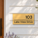 Brushed gold house number and street name sign with a brushed metal look, The Noosa, reading 103 Lake View Drive in black lettering, on white weatherboards beside a timber front door, with lavender and an olive tree.
