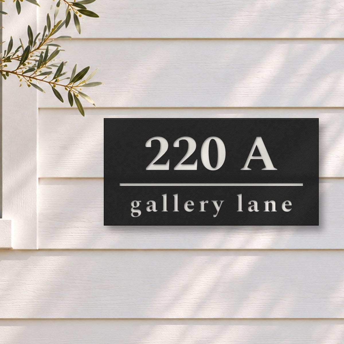 Matte black house number and street name sign, The Noosa, reading 220A Gallery Lane in white lettering, on white weatherboards beside a black-framed window and an olive tree.