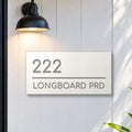 Brushed silver house number and street name sign with a brushed metal look, The Noosa, white face reading 222 Longboard Prd in silver lettering, on white cladding under a black wall light, beside a black front door.