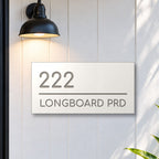 Brushed silver house number and street name sign with a brushed metal look, The Noosa, white face reading 222 Longboard Prd in silver lettering, on white cladding under a black wall light, beside a black front door.