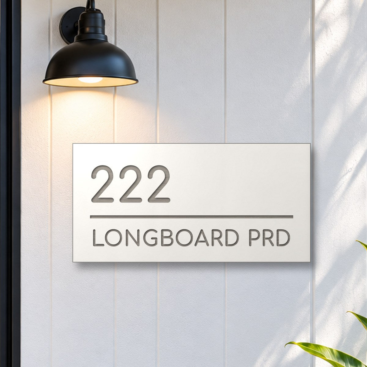 Brushed silver house number and street name sign with a brushed metal look, The Noosa, white face reading 222 Longboard Prd in silver lettering, on white cladding under a black wall light, beside a black front door.