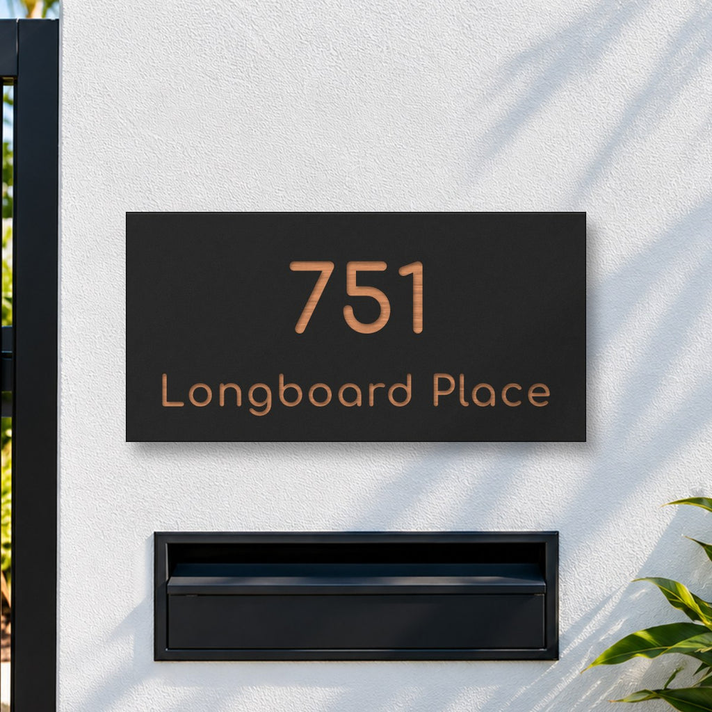 Brushed copper house number and street name sign with a brushed metal look, The Peregian, black face reading 751 Longboard Place in copper lettering, on a white rendered fence above a black letterbox.