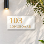 Brushed gold house number and street name sign with a brushed metal look, The Peregian, white face reading 103 Longboard in gold lettering, on a white rendered wall under a black wall light, beside an olive tree.