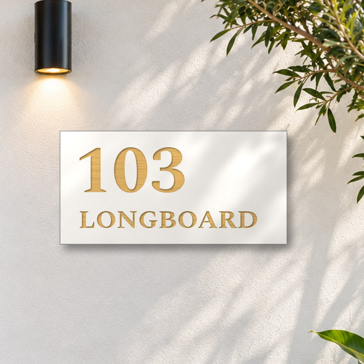 Brushed gold house number and street name sign with a brushed metal look, The Peregian, white face reading 103 Longboard in gold lettering, on a white rendered wall under a black wall light, beside an olive tree.