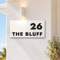 Matte white house number and street name sign, The Peregian, reading 26 The Bluff in black lettering, on a white rendered wall under a wall light, with the ocean behind.