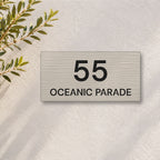 Brushed silver house number and street name sign with a brushed metal look, The Peregian, reading 55 Oceanic Parade in black lettering, on a white rendered wall with olive branches.