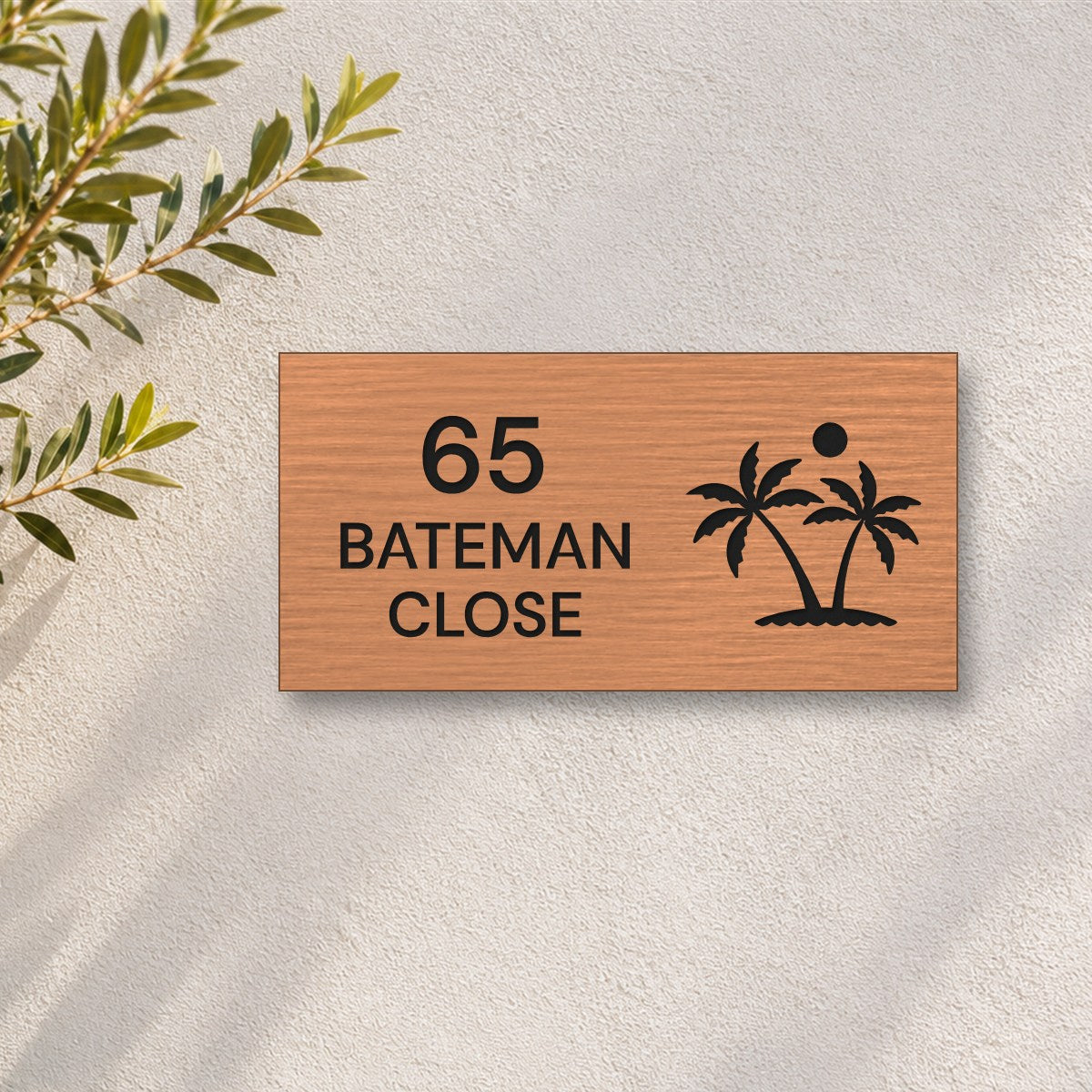 Brushed copper house number and street name sign with a brushed metal look, The Yaroomba, reading 65 Bateman Close in black lettering, with palm trees and a sun, on a white rendered wall with olive branches.